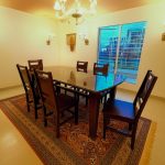 A bright dining room features a rectangular dark wood dining table with a clear glass top, surrounded by six matching dark wood chairs with slatted backs. The floor is light-colored tile, and the walls are white.