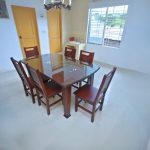 A bright dining room features a rectangular dark wood dining table with a clear glass top, surrounded by six matching dark wood chairs with slatted backs. The floor is light-colored tile, and the walls are white.