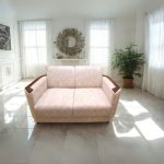 Beige two-seater fabric sofa with wooden armrests placed in a bright, modern living room with marble flooring, large windows with sheer white curtains, and a decorative mirror on the back wall.