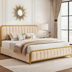 Elegant modern upholstered bed with beige padding and gold metal frame in a stylish bedroom with sunburst mirror, bedside lamp, and floor-to-ceiling curtains.