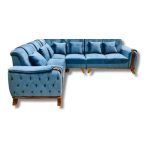 Modern living room featuring a large L-shaped blue fabric sectional sofa with wooden accents, set on a cream geometric rug. The room includes a wooden coffee table, side table with a plant, framed wall art, tall windows with blue curtains, and soft ambient lighting creating a cozy, elegant atmosphere.