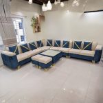 Luxurious L-shaped sectional sofa in blue and beige velvet upholstery, featuring gold metal legs and matching ottomans, set in a modern living room with bright lighting and elegant décor.