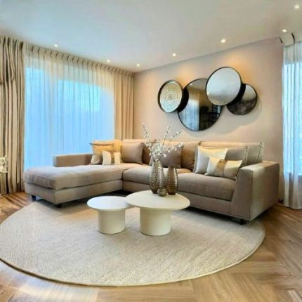 Modern beige L-shaped sectional sofa in a cozy living room with large windows, sheer curtains, circular wall mirrors, and two round white coffee tables on a textured rug.