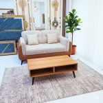 A modern living room setup featuring a two-seater beige and brown sofa with two matching cushions, placed on a textured rug. In front of the sofa is a light wooden coffee table with open storage shelves, a tall green indoor plant in a terracotta pot, and elegant gold-accented mirrors in the background with natural light streaming through sheer white curtains.