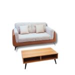 A modern living room setup featuring a two-seater beige and brown sofa with two matching cushions, placed on a textured rug. In front of the sofa is a light wooden coffee table with open storage shelves, a tall green indoor plant in a terracotta pot, and elegant gold-accented mirrors in the background with natural light streaming through sheer white curtains.