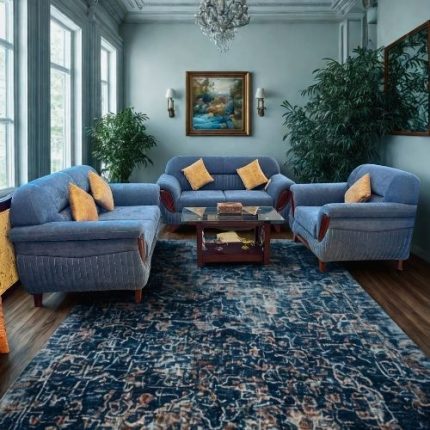 A stylish blue sofa set arranged in a bright, elegant living room featuring a patterned blue rug, wooden coffee table with glass top, yellow accent pillows, tall windows, and indoor plants creating a cozy and luxurious atmosphere.